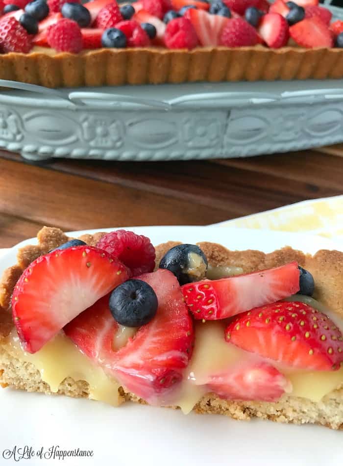 A slice of the lemon tart topped with berries on a white plate. The rest of the tart is in the background. 
