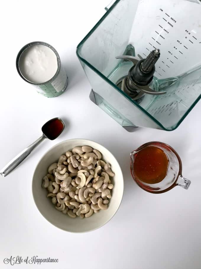 A can of coconut milk, vanilla extract, bowl of cashews, and cup of honey surrounding a blender. 