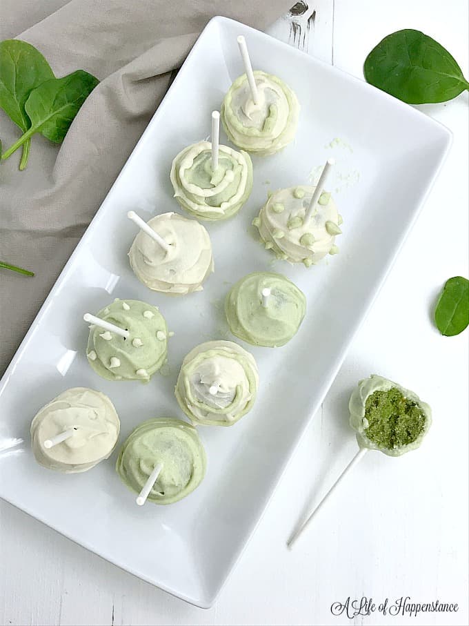 Green and white almond flour vanilla cake pops on a rectangular, white dish. A half eaten cake pop on the table next to the dish.  