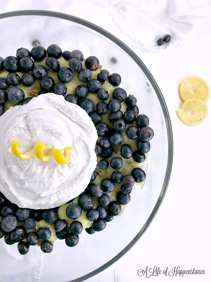 An overhead shot of lemon curd, blueberries, and whipped coconut cream topped with a curled lemon rind in a glass trifle dish. 