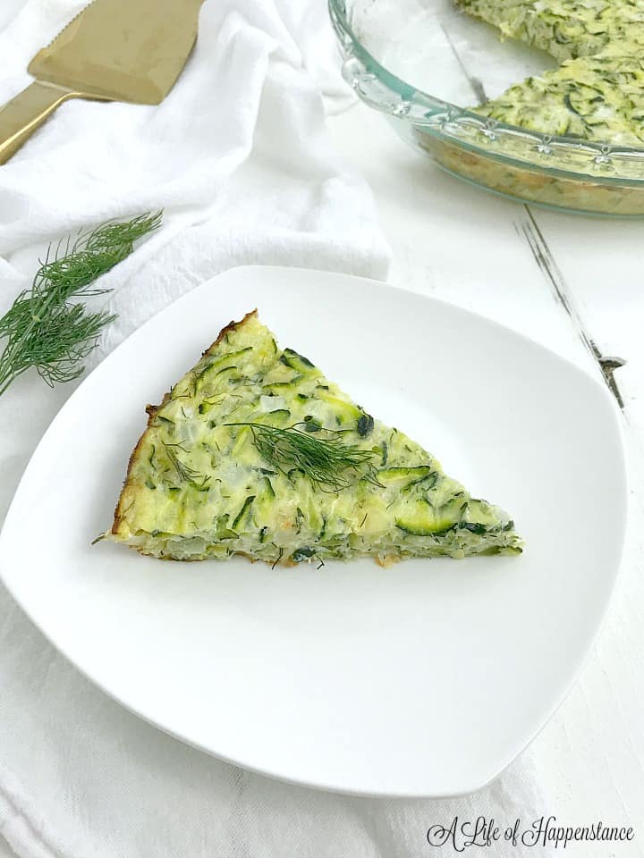 A slice of quiche on a white plate with the rest of the zucchini quiche in the background. 