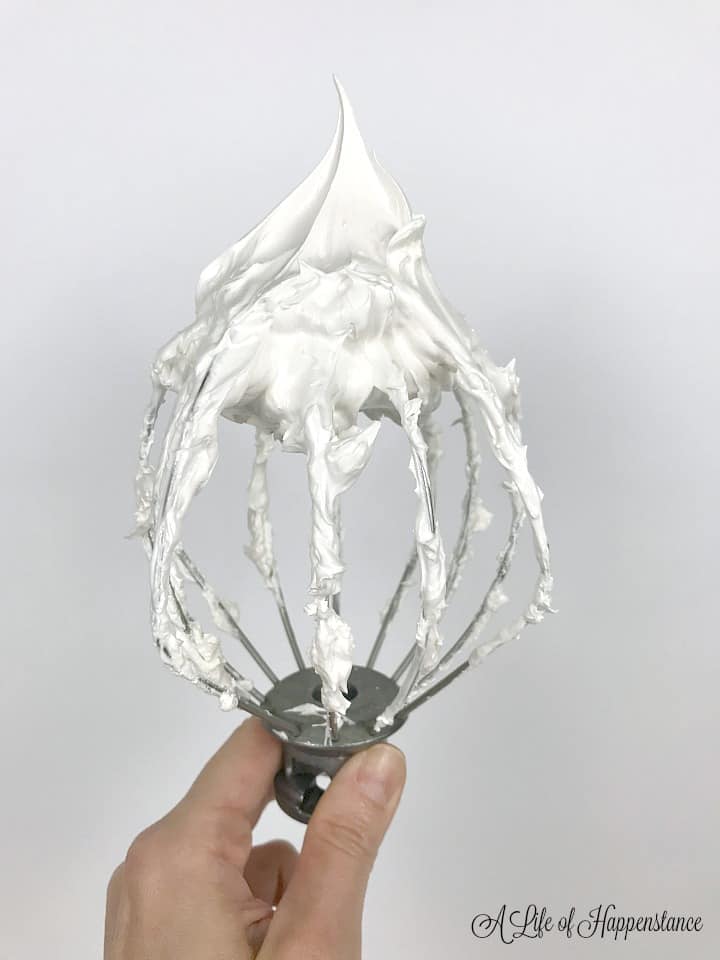 A hand holding a whisk filled with white Italian meringue. 