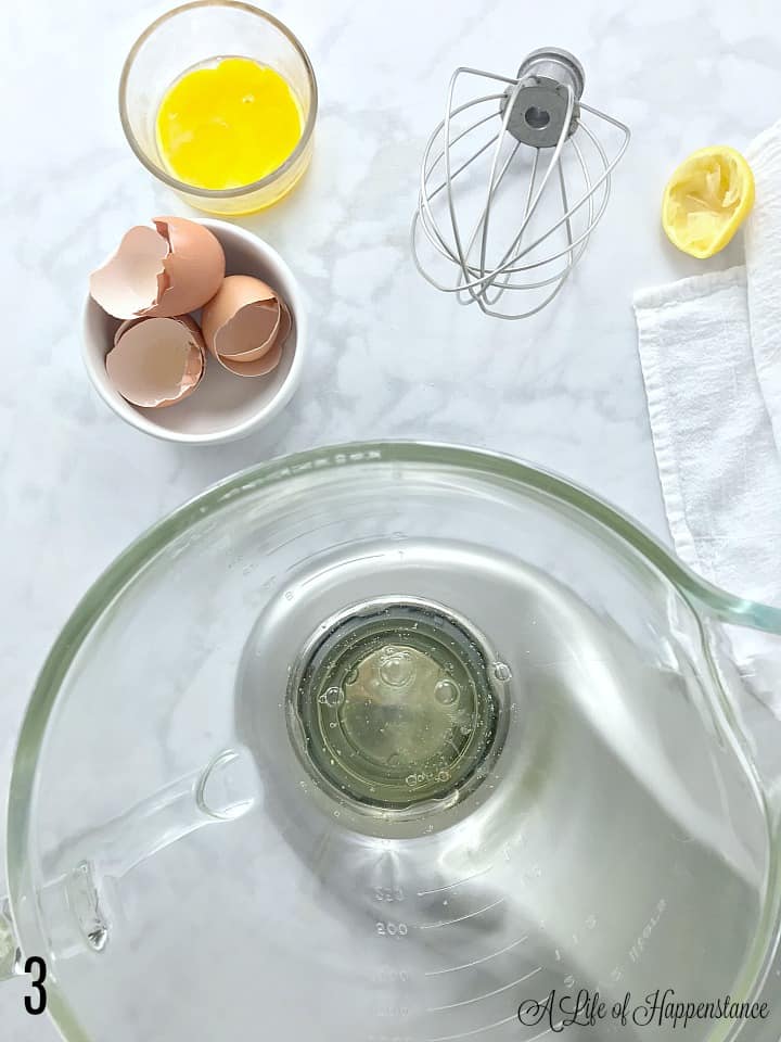 A mixing bowl filled with egg whites, a whisk, egg shells, and half of a lemon on a marble table. 