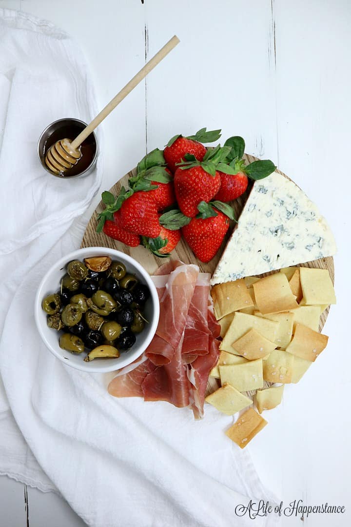  A small charcuterie board filled with almond flour crackers, strawberries, prosciutto, olives, and a wedge of cheese. 