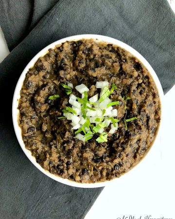 A bowlful of homemade refried black beans garnished with onion and cilantro in a white bowl on a grey kitchen towel.