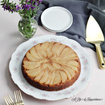 The upside-down paleo pear cake on a white plate surrounded by purple flowers, white plates, and a gray kitchen towel.