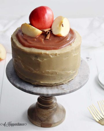 The apple cider layer cake on a cake stand with sliced apples and cinnamon sticks on top.